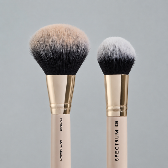 Two makeup brushes with cream-colored handles, gold ferrules, and black bristles, shown against a solid gray background. One brush is labeled 'POWDER' and the other 'COMPLEXION'.