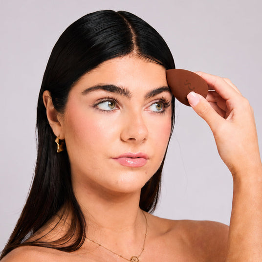 A woman with long dark hair applies a brown makeup product, likely a brow pomade or concealer, using a small applicator. She is holding a compact mirror in her other hand, set against a plain light gray studio background.
