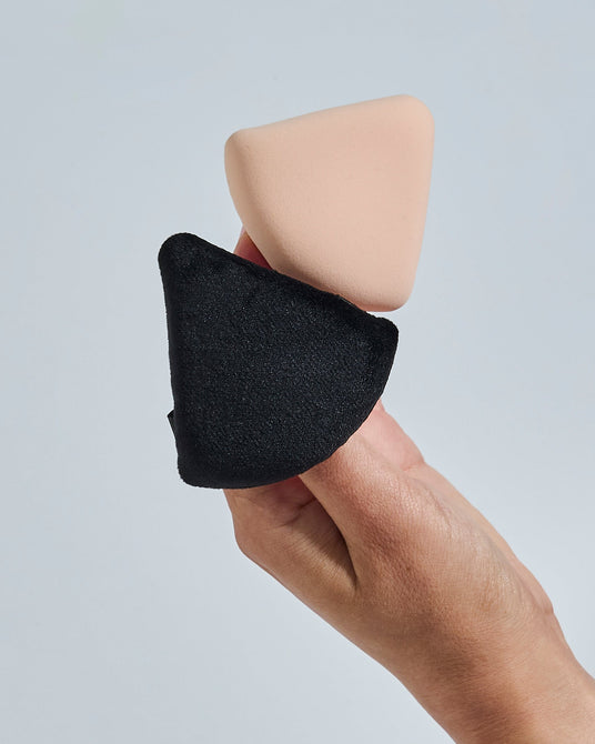 A hand holding a black fabric-covered triangular makeup sponge with a beige sponge block attached, against a light gray background.