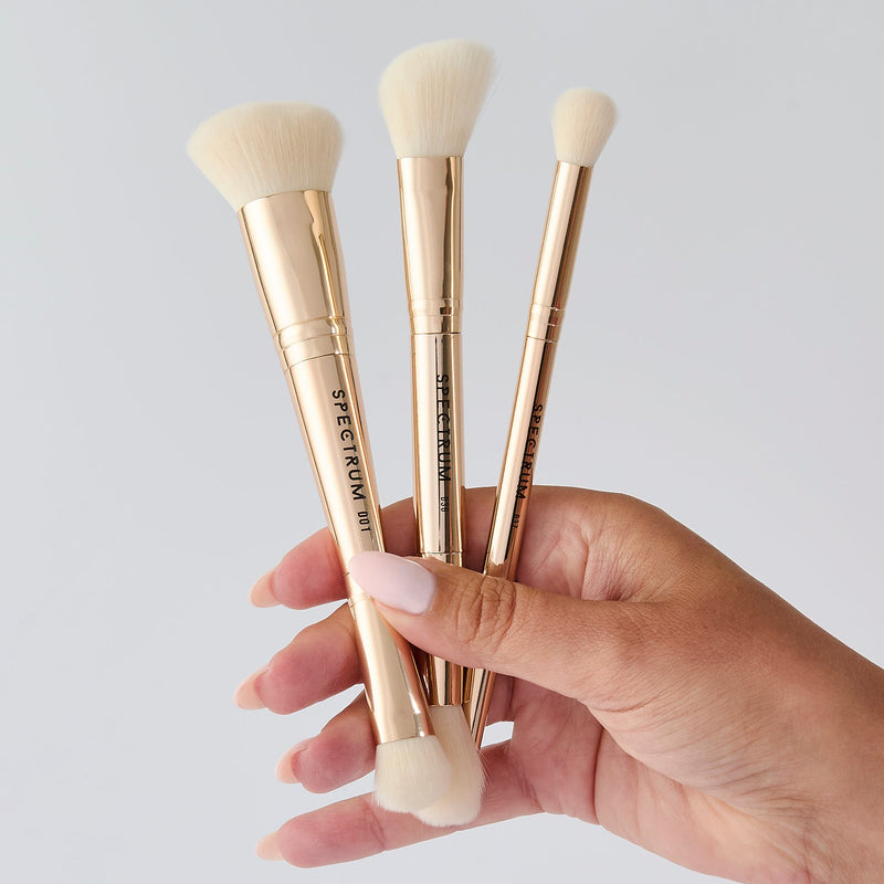 Three makeup brushes with pale gold handles and cream bristles are held in a person's hand against a light gray background.