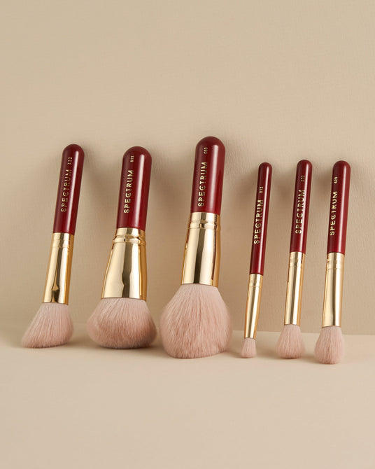 Six makeup brushes with deep red handles, gold ferrules, and pale pink bristles, arranged upright on a warm beige background. The brand name 'SPECTRUM' is printed vertically on the handles.