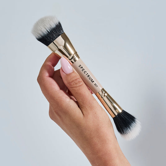 A hand with light pink manicured nails holds a makeup brush with black and white bristles and a gold handle against a light gray background. The brand name 'SPECTRUM' is visible on the handle.