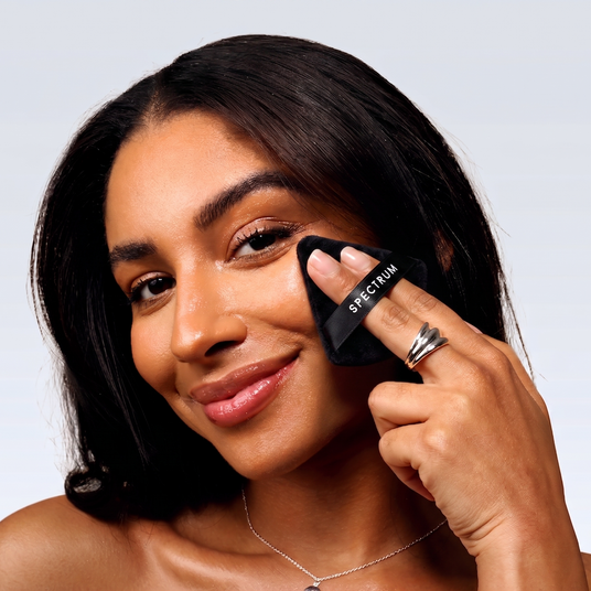 A close-up of a woman with dark hair applying or blending with black cosmetic puff with the brand name 'SPECTRUM' visible on it, against a clean light gray background.